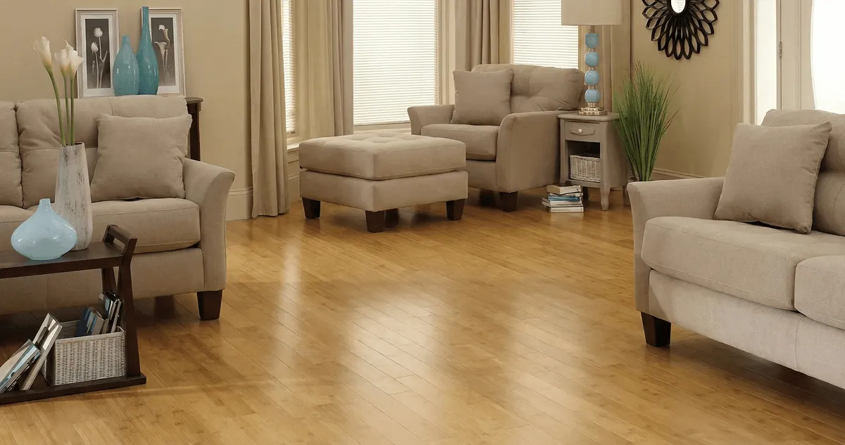 A living room with professionally installed bamboo flooring, beige sofas, a matching ottoman, a side table with a blue lamp, vases, framed pictures, and large windows with blinds and curtains in Nashville, TN